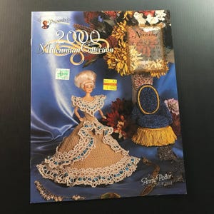 May include: A vintage craft pattern book titled "2000 Millennium Collection" featuring a doll in a crocheted gown. The cover has a blue background with floral arrangements and a small chair. The doll's dress is beige with white and blue trim.