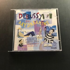 May include: A CD case featuring the album "Debussy for Daydreaming." The cover art includes illustrations of people in various relaxed poses, with text that reads "Music to Caress Your Innermost Thoughts." The Philips logo is visible.