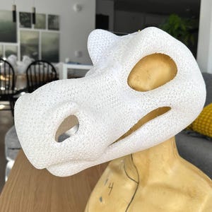May include: A white, textured, dinosaur-like mask with large eye and mouth openings. The mask is mounted on a tan mannequin head. The mask is likely for cosplay or costume use.