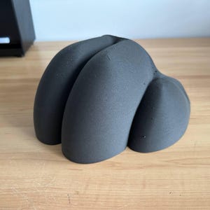 May include: A dark gray, abstract sculpture with a textured surface. The form has three rounded, organic shapes that are connected. The sculpture is placed on a light brown wooden surface.