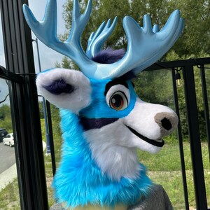 Pu&ograve; includere: Una testa di animale di peluche blu e bianca con grandi corna blu. La testa &egrave; fatta di tessuto peluche e ha un'espressione amichevole. L'animale indossa una camicia grigia.