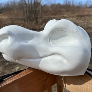 May include: White unpainted furry animal head base for a costume. The head is made of foam and has a closed mouth with a defined nose and eye sockets.