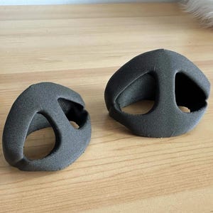 May include: Two gray, foam-like ear cushions, designed to fit over the ears. The cushions have a curved shape with a large opening in the center and a smaller opening on the side. They are placed on a light brown wooden surface.
