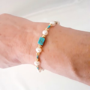 May include: A delicate bracelet featuring white pearls, gold beads, and small turquoise accents. The focal point is a rectangular turquoise stone. The bracelet is strung on a gold chain, creating an elegant and refined piece of jewelry.