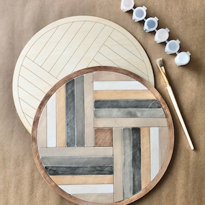 May include: A round wooden tray with a geometric pattern painted in shades of grey, brown, and white. The tray is unfinished and ready to be painted. A set of paint pots in various shades of grey and brown are included.