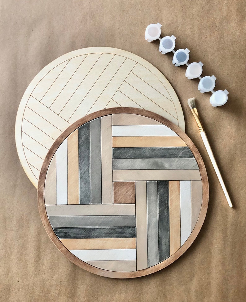 May include: A round wooden tray with a geometric pattern painted in shades of grey, brown, and white. The tray is unfinished and ready to be painted. A set of paint pots in various shades of grey and brown are included.