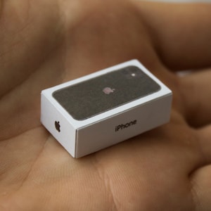 Può includere: Una scatola bianca in miniatura con il logo Apple e la scritta "iPhone" stampata su di essa. La scatola è aperta, rivelando un iPhone nero all'interno.