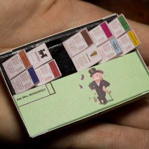 May include: Miniature Monopoly game set inside a matchbox. The game includes property cards, a green game board with the Monopoly man, and the text "Ask Mrs. Monopoly". The set is held in a person's hand.