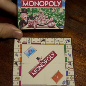 May include: A miniature Monopoly board game set, featuring a game box with the Monopoly logo and illustrations of game pieces. The game board is also visible, with property spaces and the Monopoly logo.