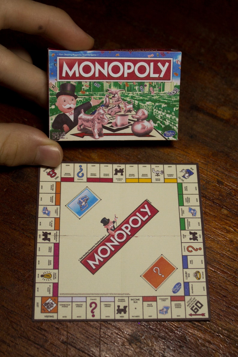 May include: A miniature Monopoly board game set, featuring a game box with the Monopoly logo and illustrations of game pieces. The game board is also visible, with property spaces and the Monopoly logo.