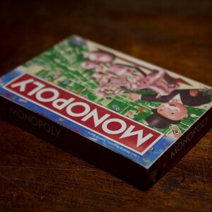 May include: A Monopoly board game box, featuring the game's logo in red and white on a blue background. The box displays illustrations of the game's characters and board. The box is resting on a dark wooden surface.