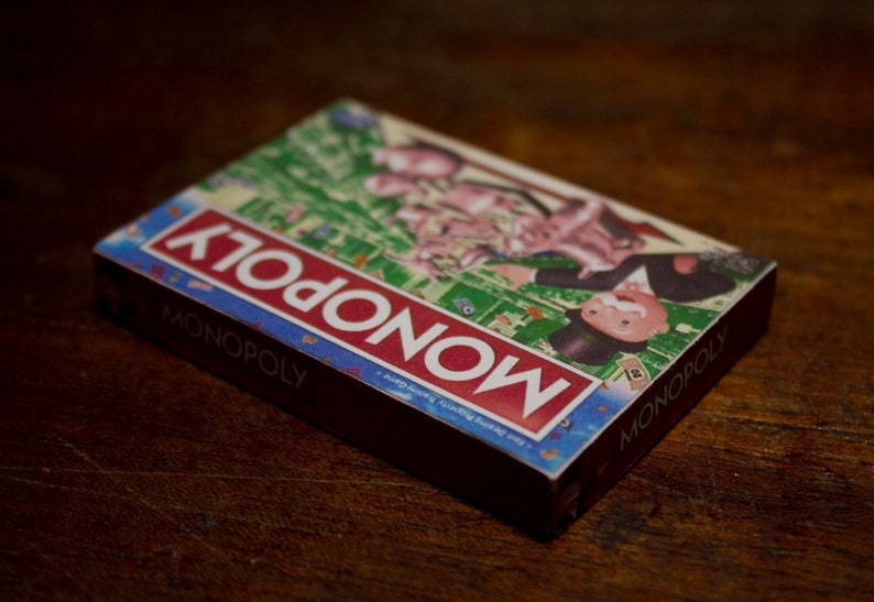 May include: A Monopoly board game box, featuring the game's logo in red and white on a blue background. The box displays illustrations of the game's characters and board. The box is resting on a dark wooden surface.