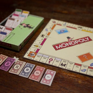 May include: A Monopoly board game set with a green box, game board, play money, and property cards. The board game is on a wooden surface. The Monopoly logo is visible on the board and box.