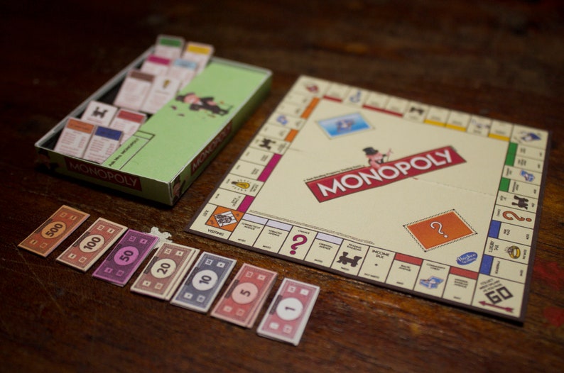 May include: A Monopoly board game set with a green box, game board, play money, and property cards. The board game is on a wooden surface. The Monopoly logo is visible on the board and box.