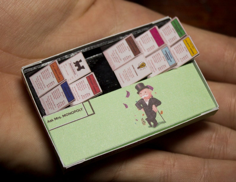 May include: Miniature Monopoly game set inside a matchbox. The game includes property cards, a green game board with the Monopoly man, and the text "Ask Mrs. Monopoly". The set is held in a person's hand.