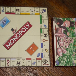 May include: A Monopoly board game set, featuring the game board and the box. The board is beige with property spaces and the word "MONOPOLY" in red. The box shows the game's mascot and the word "MONOPOLY".