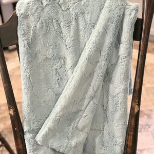 May include: A soft, light blue plush blanket draped over a wooden chair. The blanket has a textured, fur-like appearance, with visible folds and a cozy aesthetic. The chair has a dark wood finish, with visible wear and tear.