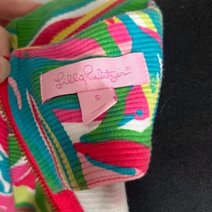 May include: A pink, green, and blue patterned zippered top with a Lilly Pulitzer label. The label is pink and says "Lilly Pulitzer" in white letters. The size tag is attached to the label and says "S" in white letters.