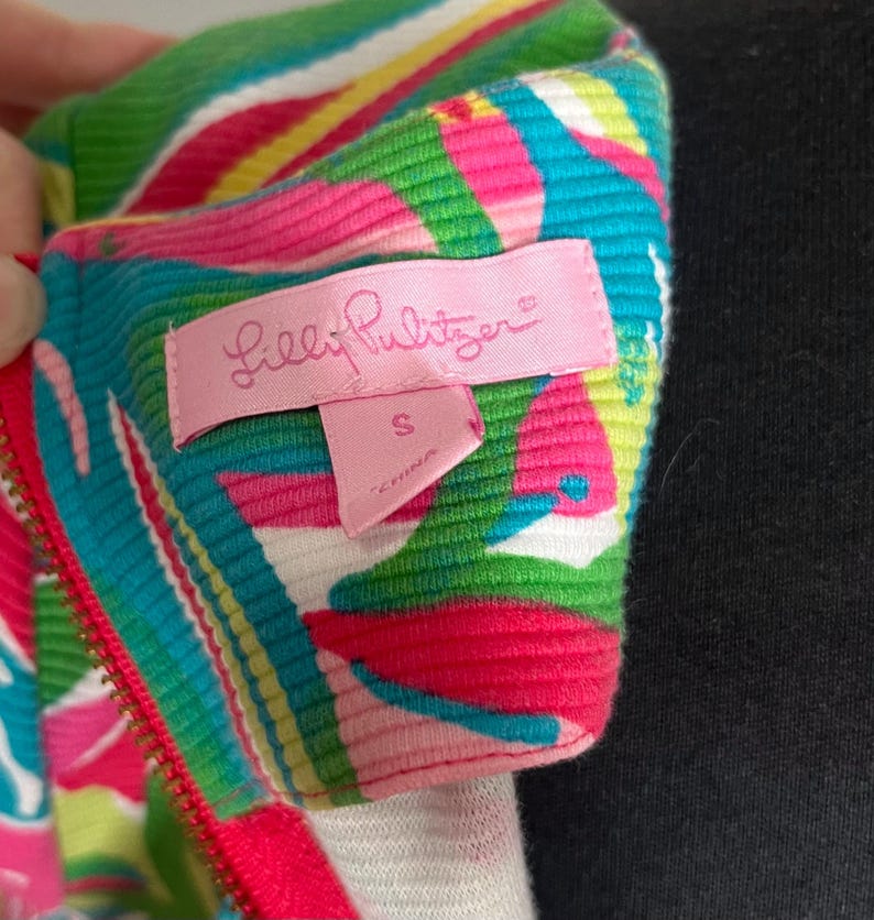 May include: A pink, green, and blue patterned zippered top with a Lilly Pulitzer label. The label is pink and says "Lilly Pulitzer" in white letters. The size tag is attached to the label and says "S" in white letters.