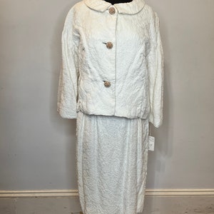 May include: White textured skirt suit with a peter pan collar jacket and pearl-like buttons.