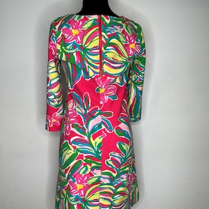 May include: A pink dress with a floral print featuring pink, green, and yellow flowers and leaves. The dress has a boat neck and three-quarter sleeves. It has a zipper closure in the back.