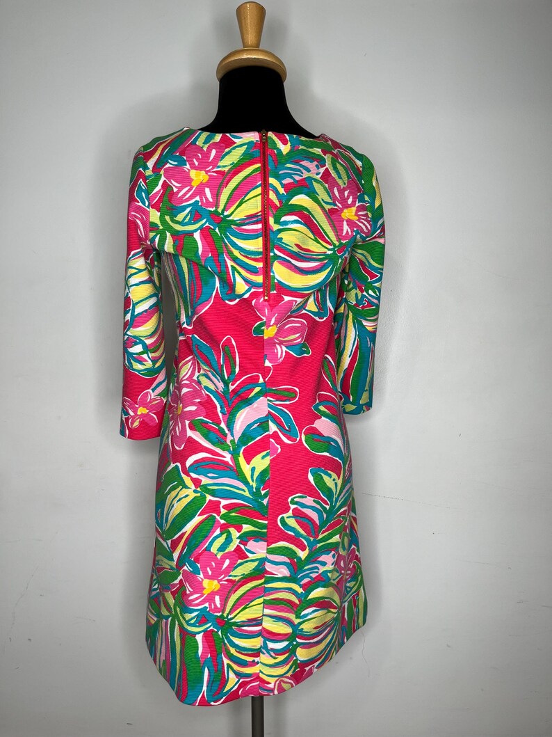 May include: A pink dress with a floral print featuring pink, green, and yellow flowers and leaves. The dress has a boat neck and three-quarter sleeves. It has a zipper closure in the back.