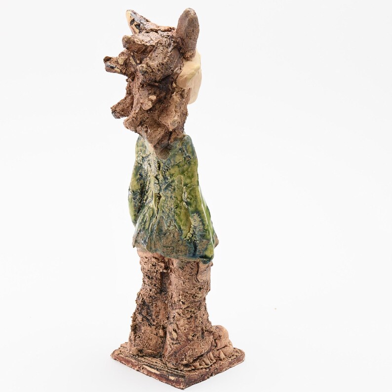 Puede incluir: Una escultura de cer&aacute;mica hecha a mano de una figura con una apariencia texturizada y &aacute;spera. La figura tiene una chaqueta verde y marr&oacute;n, con una cara de color claro y una cabeza texturizada. La escultura est&aacute; montada sobre una base cuadrada.