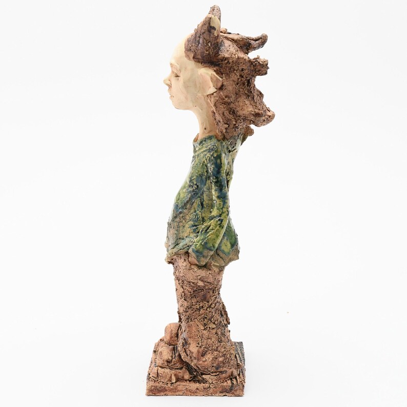 Puede incluir: Escultura de cer&aacute;mica de una figura de perfil, con rostro claro y cabello casta&ntilde;o texturizado. La figura viste una parte superior texturizada verde y azul y pantalones marrones, de pie sobre una base marr&oacute;n. La obra de arte tiene una est&eacute;tica r&uacute;stica y artesanal.
