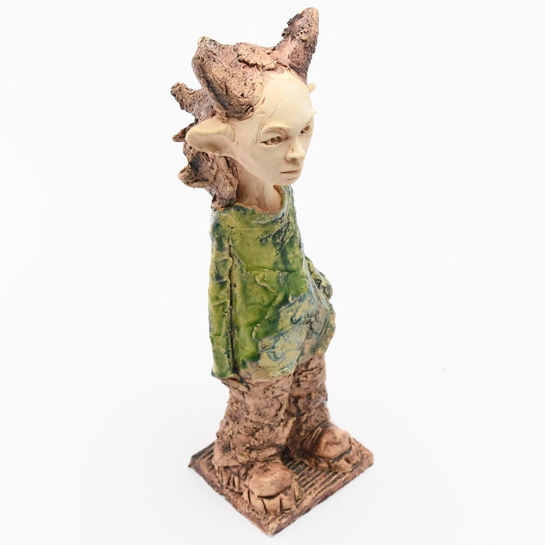 Puede incluir: Una escultura de cer&aacute;mica de una figura humanoide con una expresi&oacute;n serena. La figura tiene una cara p&aacute;lida, orejas puntiagudas y un tocado marr&oacute;n texturizado. Viste una t&uacute;nica verde y pantalones marrones texturizados. La escultura se alza sobre una base cuadrada.