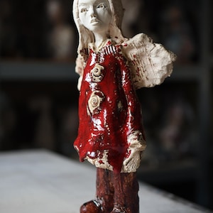 May include: A ceramic figurine of a figure with a white face and hair, wearing a glossy red coat with three circular textured buttons. The figure has large, rough-textured wings, dark brown trousers, and brown boots, standing on a square base.