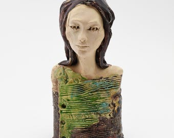 May include: A ceramic bust of a woman with long brown hair and a serene expression. The figure's upper body is adorned with a textured green and brown garment. The artwork has a handcrafted appearance.