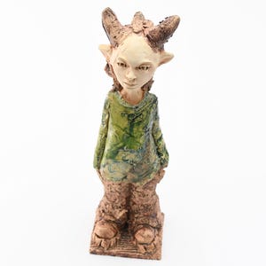 Puede incluir: Figura de cer&aacute;mica de una criatura con cuernos, orejas puntiagudas y una camisa verde. La figura tiene una expresi&oacute;n neutra y se encuentra sobre una base texturizada. Los colores son tonos tierra de marr&oacute;n, verde y beige.