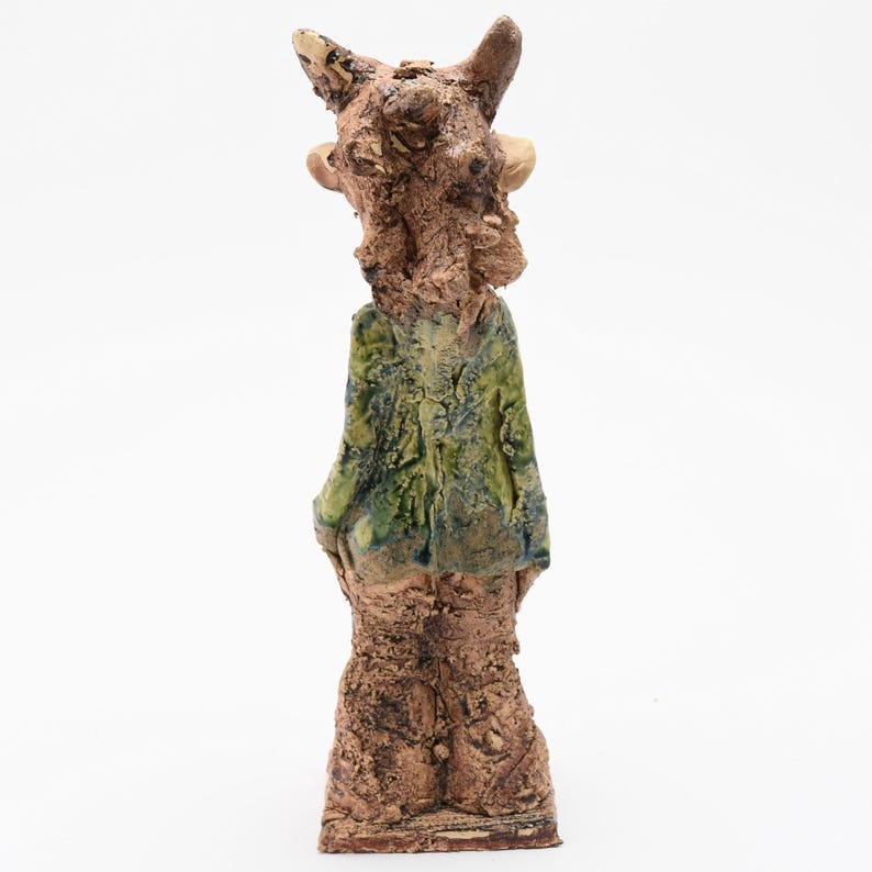 Puede incluir: Escultura de cer&aacute;mica de una figura con cuerpo y cabeza marrones texturizados. La figura lleva una chaqueta esmaltada verde y azul. La escultura tiene un aspecto rugoso y org&aacute;nico y se apoya sobre una base rectangular.