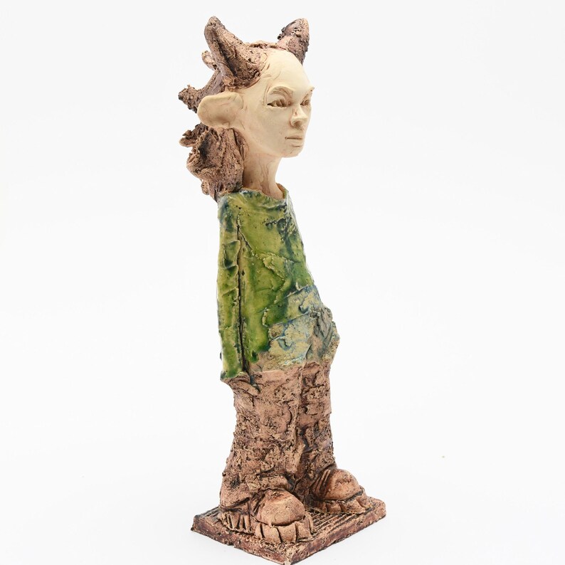 Puede incluir: Escultura de cer&aacute;mica de una figura humanoide con tez clara, que lleva una camisa verde y pantalones marrones texturizados. La figura tiene orejas puntiagudas y cuernos, de pie sobre una base cuadrada. La escultura est&aacute; sobre un fondo blanco.