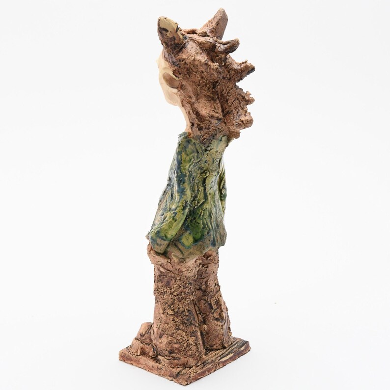 Puede incluir: Escultura de cer&aacute;mica hecha a mano de una figura con cuerpo y cabeza marrones texturizados. La figura lleva una camisa verde y azul. La escultura tiene un dise&ntilde;o abstracto &uacute;nico y est&aacute; sobre un fondo blanco.