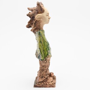 Puede incluir: Escultura de cer&aacute;mica de una figura con un peinado texturizado y puntiagudo y una expresi&oacute;n facial serena. La figura lleva una prenda verde y se apoya sobre una base texturizada. La escultura es predominantemente marr&oacute;n y verde.