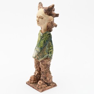 Puede incluir: Una escultura de cer&aacute;mica de una figura con tez clara, vestida con un atuendo verde y marr&oacute;n. La figura tiene el pelo puntiagudo y pies grandes. La escultura est&aacute; sobre una base cuadrada.