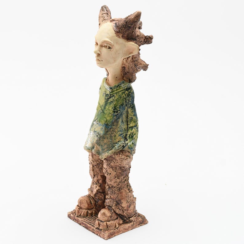 Puede incluir: Una escultura de cer&aacute;mica de una figura con tez clara, vestida con un atuendo verde y marr&oacute;n. La figura tiene el pelo puntiagudo y pies grandes. La escultura est&aacute; sobre una base cuadrada.