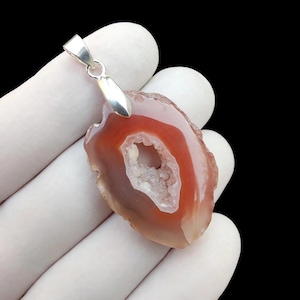 May include: A pendant featuring a polished, translucent reddish-orange agate slice with a central geode cavity filled with small, sparkling white crystals. The stone is secured by a silver-toned metal bail at the top for suspension.