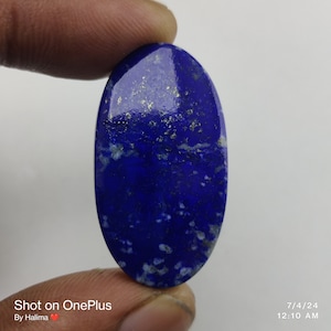 Op de afbeelding: Een gepolijste, ovaalvormige lapis lazuli edelsteen. De diepblauwe steen heeft gouden vlekken en een glad, reflecterend oppervlak. De edelsteen wordt tussen vingers gehouden tegen een witte achtergrond. De afbeelding bevat tekst en de datum.