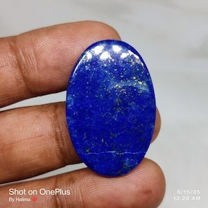 Op de afbeelding: Een ovale, gepolijste lapis lazuli edelsteen. De diepblauwe steen heeft gouden vlekken en een glad, glanzend oppervlak. De afbeelding bevat de tekst "Shot on OnePlus" en de datum en tijd "8/15/25 12:29 AM".