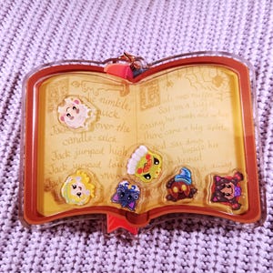 May include: A clear acrylic book-shaped charm with a brown border, featuring a yellow interior with text and cartoon character designs. The charm has a red ribbon and a gold-colored clasp.