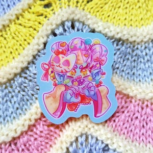 May include: A vibrant sticker showcasing a fantastical character with a cat mask, unicorn horn, and multiple eyes. The design uses pink, blue, and yellow tones, in a cartoon style. The sticker is placed on a backdrop of pastel-coloured knitted fabric.