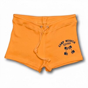 Puede incluir: Pantalones cortos de color naranja brillante con cintura de cord&oacute;n y un gr&aacute;fico azul oscuro en la parte inferior derecha. El gr&aacute;fico muestra el texto CAMP MUSCLE BODYWEAR sobre una ilustraci&oacute;n de dos mancuernas inclinadas en forma de V.