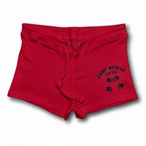 Puede incluir: Pantalones cortos deportivos de color rojo brillante con cintura de cord&oacute;n y un gr&aacute;fico azul oscuro en la pierna derecha. El gr&aacute;fico muestra el texto CAMP MUSCLE BODYWEAR arqueado sobre una ilustraci&oacute;n de dos mancuernas cruzadas con patrones de bandera estadounidense.