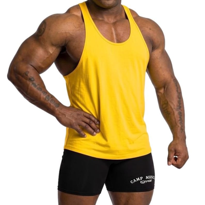 May include: A bright yellow sleeveless tank top with a racerback design paired with black athletic shorts. The shorts feature the white text "CAMP MUSCLE" printed on the lower left leg, accompanied by a small circular logo graphic below the text.