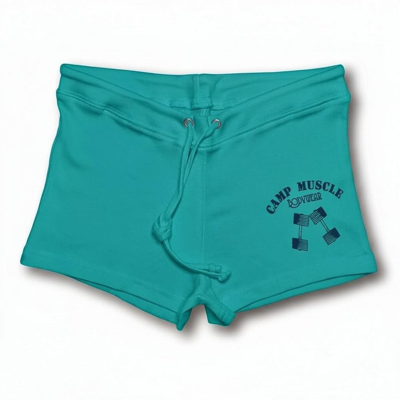 Puede incluir: Pantalones cortos de color verde azulado con cintura de cord&oacute;n y un gr&aacute;fico azul oscuro en la parte inferior derecha. El gr&aacute;fico muestra el texto CAMP MUSCLE BODYWEAR arqueado sobre una ilustraci&oacute;n de dos mancuernas en &aacute;ngulo.