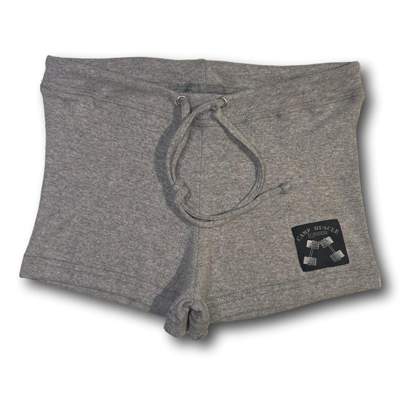 Puede incluir: Pantalones cortos de color gris jaspeado con cintura de cord&oacute;n y ojales met&aacute;licos plateados. Un parche rectangular negro est&aacute; cosido en la parte inferior derecha, mostrando el texto "CAMP MUSCLE" sobre una ilustraci&oacute;n gr&aacute;fica de dos mancuernas.