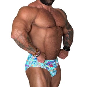 Pode incluir: Cueca de banho azul-clara com uma estampa vibrante em estilo de banda desenhada, apresentando estrelas coloridas, relâmpagos e balões de texto com palavras como "PLUNK" e "SPLASH" em letras gráficas arrojadas por toda a superfície.