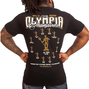 May include: Black t-shirt with gold and white text reading "Mr. Olympia Champions" above a graphic of a bodybuilder holding a trophy. The shirt lists past champions and the text "PHIL HEATH 2011-2014".